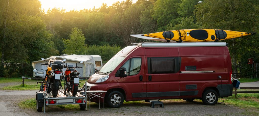 Sprinter van with kayaks on top parked in a campground next to a motorcycle trailer.